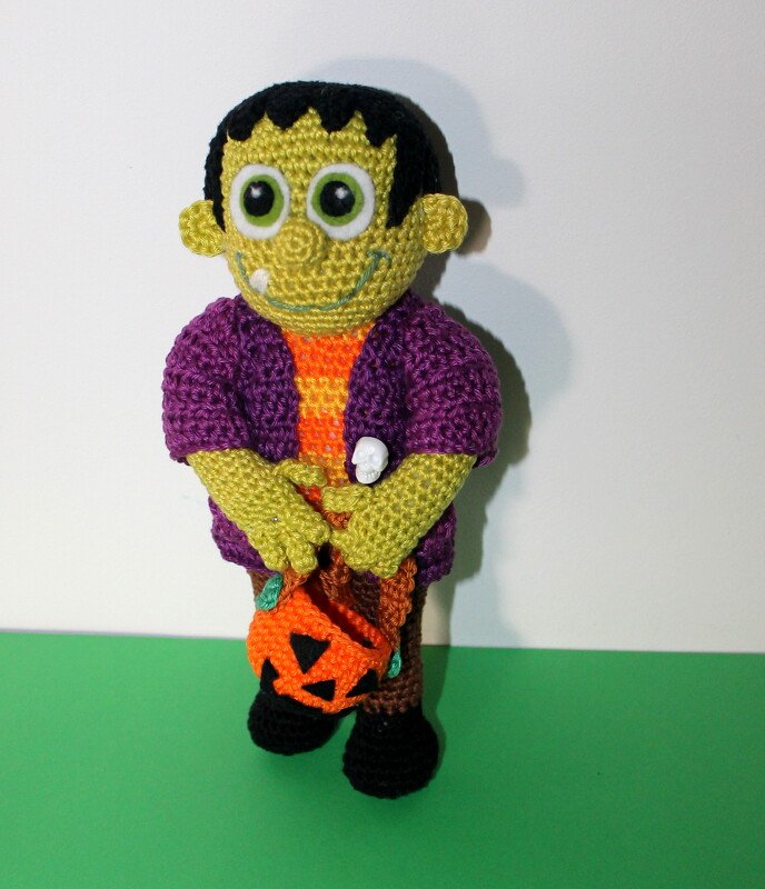 Green crochet amigurumi doll in purple jacket holding a pumpkin bag