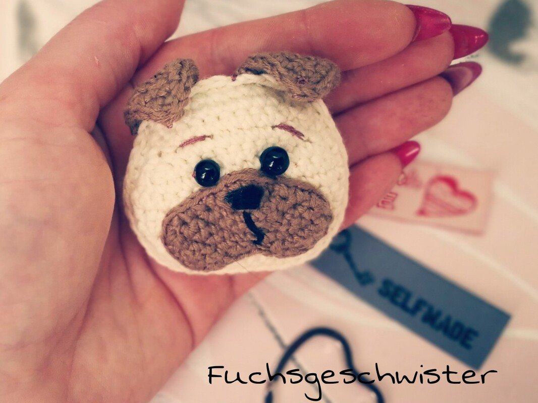 Hand hält kleinen gehäkelten Hundekopf-Amigurumi in Creme und Braun