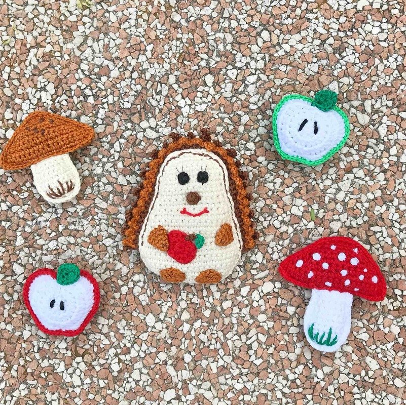 Gehäkelter Igel mit Apfel, zwei gehäkelte Pilze und zwei Apfelscheiben auf kiesigem Untergrund