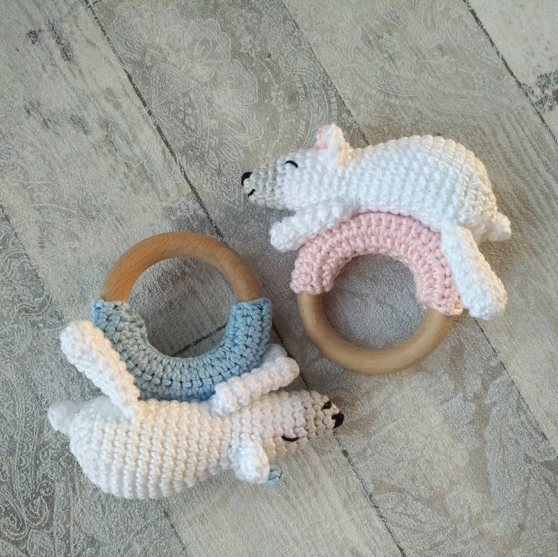 Zwei gehäkelte Babyrasseln mit weißen Tierfiguren an Holzringen, Details in Hellblau und Rosa auf hellem Holzboden.