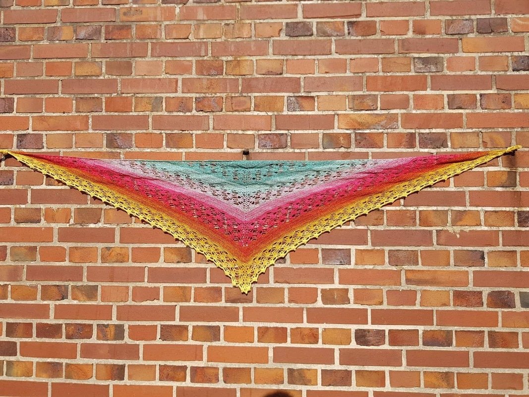 Triangle crochet shawl with yellow, pink and teal gradient lace stitches and scalloped edge hanging on a brick wall.