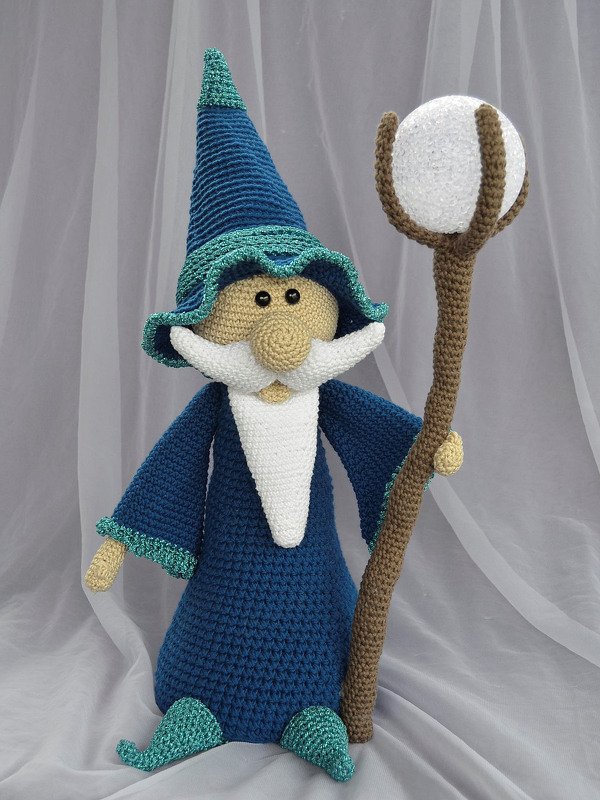 Gehäkelte Amigurumi-Zauberer-Puppe in blauem Gewand mit weißem Bart und Zauberstab mit weißer Kugel