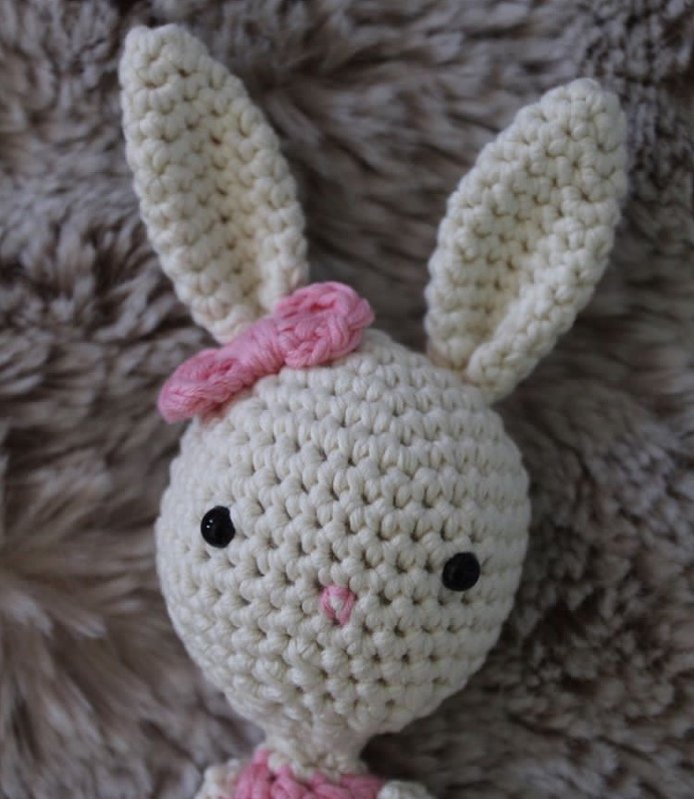 Cremeweißes Amigurumi-Häschen mit rosa Schleife und schwarzen Augen auf flauschigem Untergrund