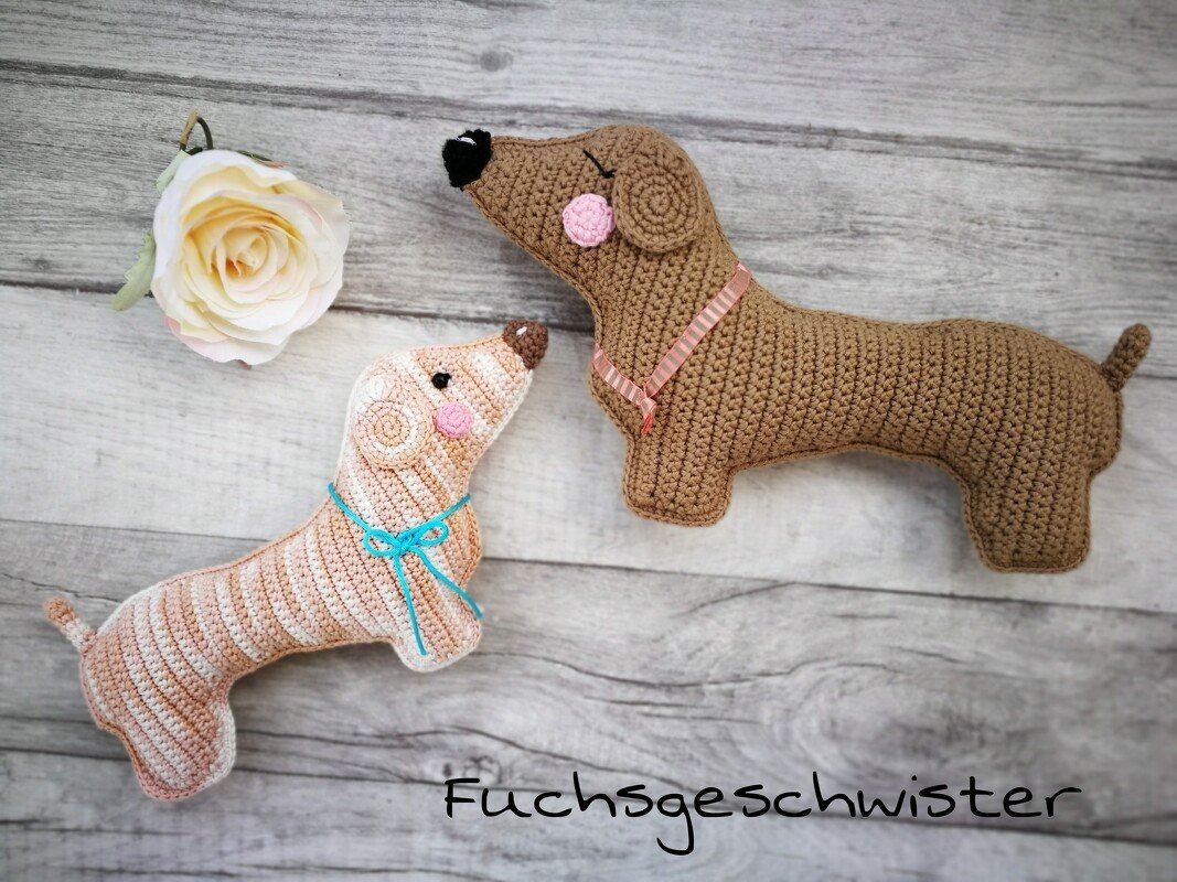 Zwei gehäkelte Dackel-Amigurumi auf hellem Holzuntergrund, einer gestreift mit blauem Band, der andere braun mit rosa Halsband
