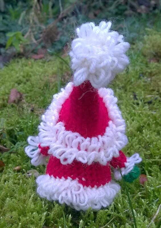Kleine gehäkelte Weihnachtsfigur in roter Zipfelmütze mit weißem Bommel auf grünem Moos