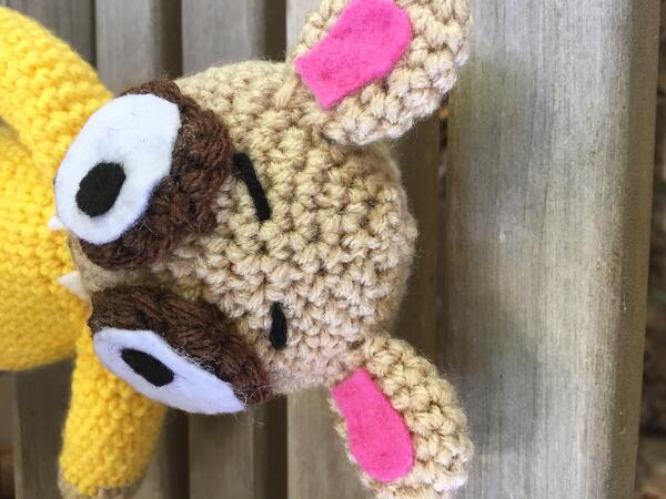Crocheted amigurumi doll head in light brown yarn with pink inner ears and felt eyes on wooden background.