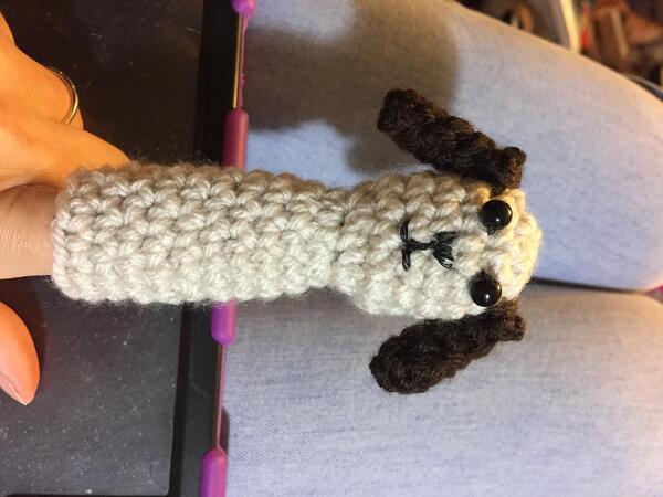 Hand holding a cream crochet dog finger puppet with dark brown ears and a purple crochet hook in view