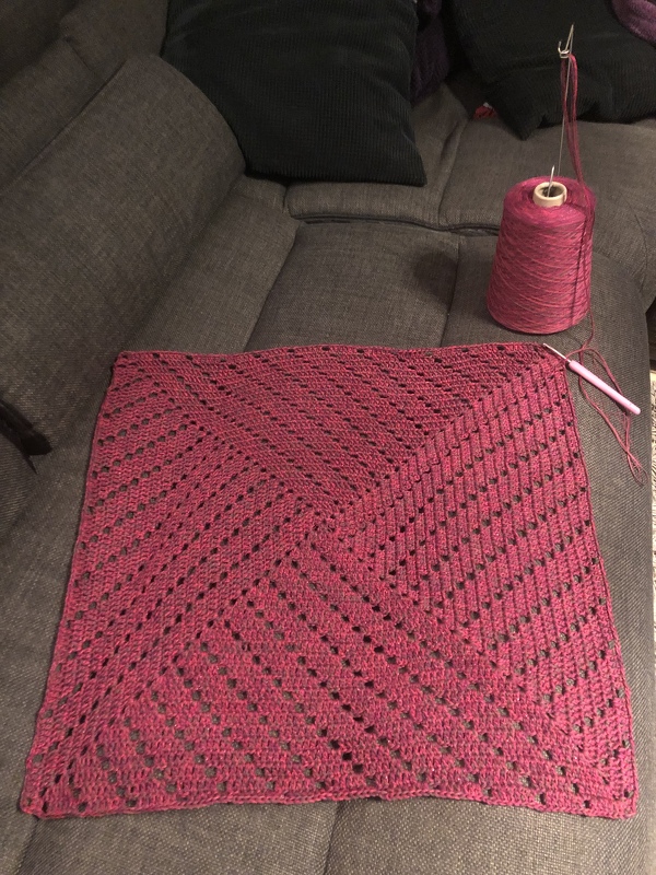 Unvollendetes gehäkeltes Quadrat in Magenta mit diagonalem Rautenmuster, Garnkone und Häkelnadel auf Sofa.