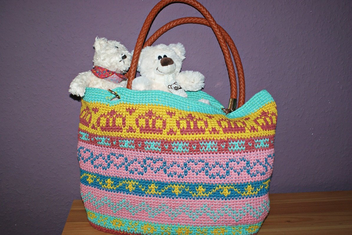 Gehäkelter bunter Shopper mit Streifenmuster und braunen Ledergriffen, zwei weiße Stofftiere darin, lila Hintergrund.
