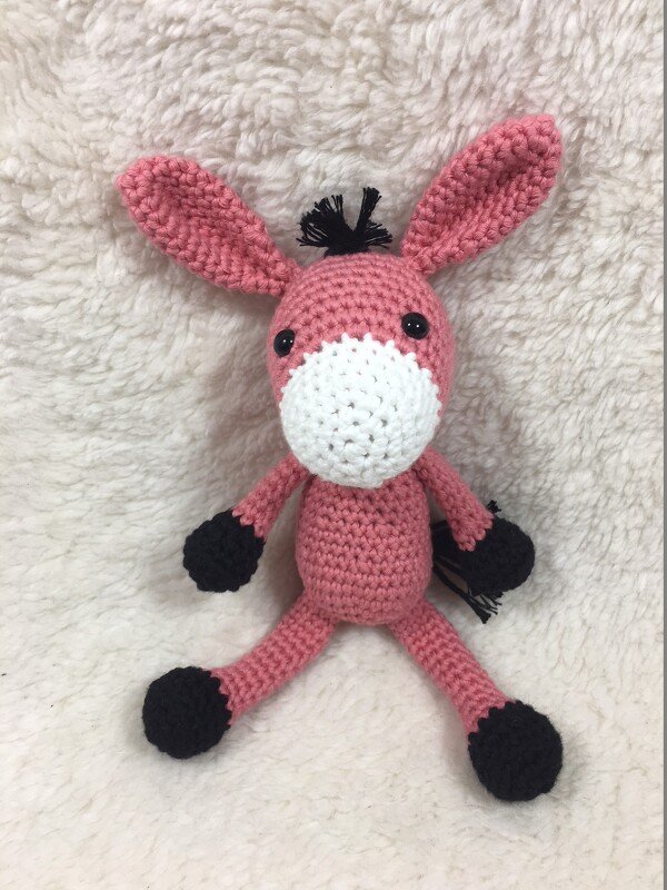Rosa gehäkelter Amigurumi‑Esel mit weißer Schnauze und schwarzen Details, sitzend auf hellem, flauschigem Stoff.