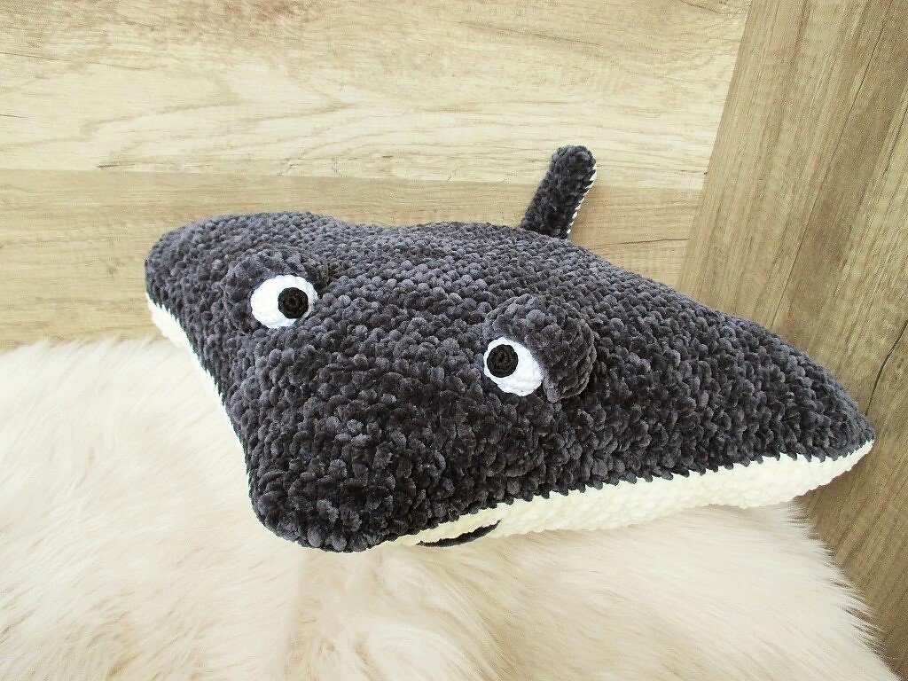 Gehäkeltes Rochen-Kissen aus dunkelgrauem Plüschgarn mit weißen Augen auf hellem Fellvorleger vor einer Holzwand.