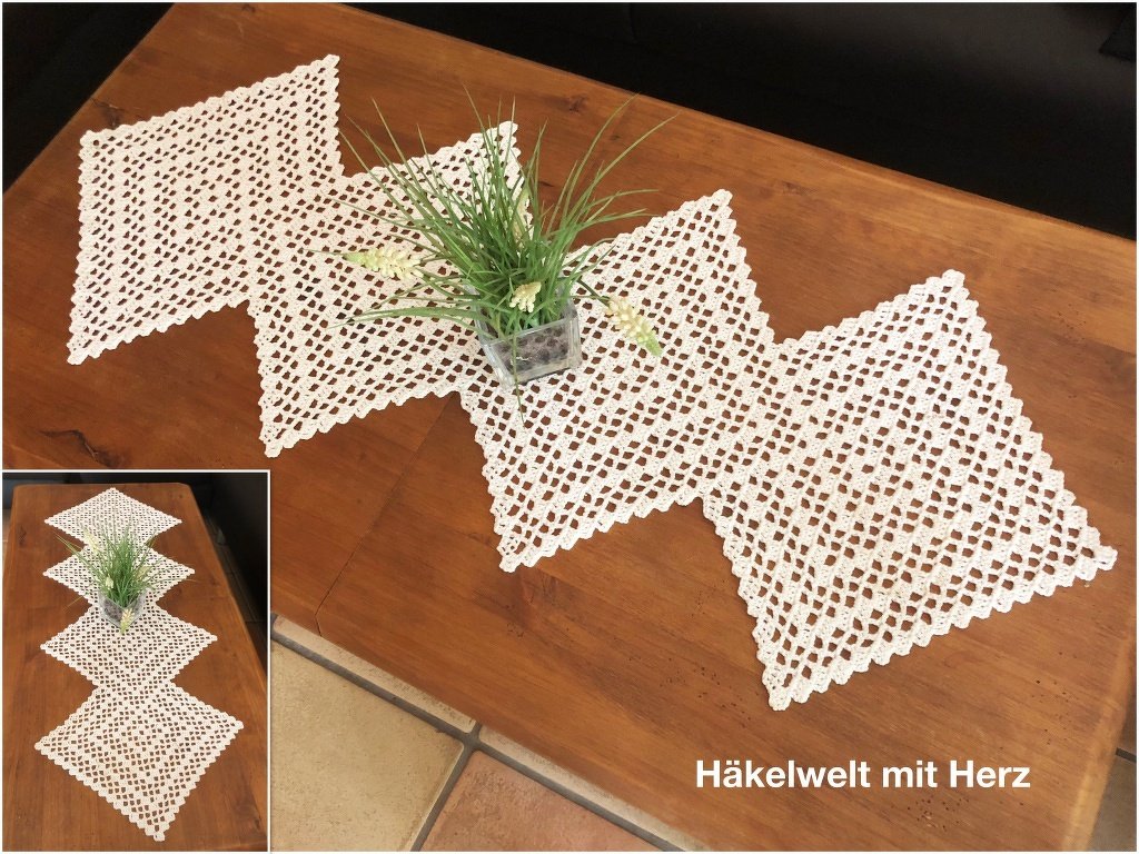 Weiß gehäkelter Tischläufer mit Rautenmuster auf braunem Holztisch, mit kleiner Topfpflanze dekoriert.