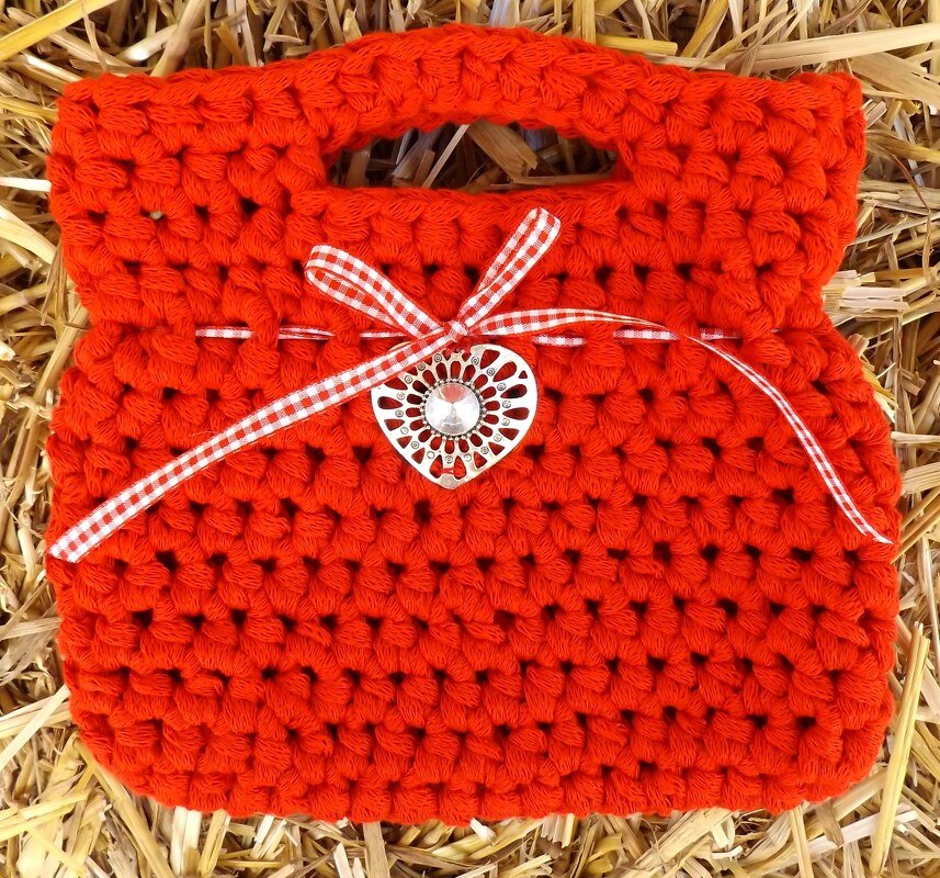 Gehäkelte Henkeltasche in kräftigem Rot mit Grifföffnung, kariertem Band und Herzanhänger auf Stroh