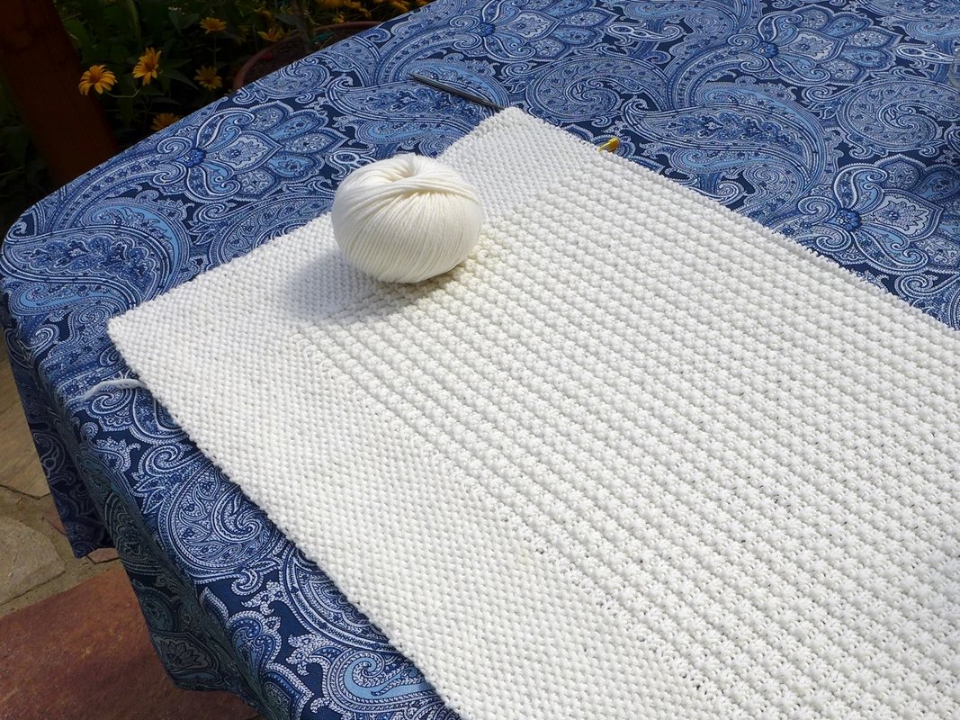 White knitted blanket or sack in progress with a ball of white yarn on a blue paisley tablecloth.