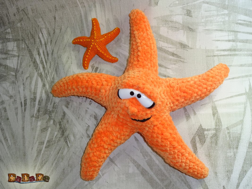 Zwei orangefarbene gehäkelte Seesterne, ein großes mit Gesicht und ein kleiner ohne Gesicht, auf hellem Stoff