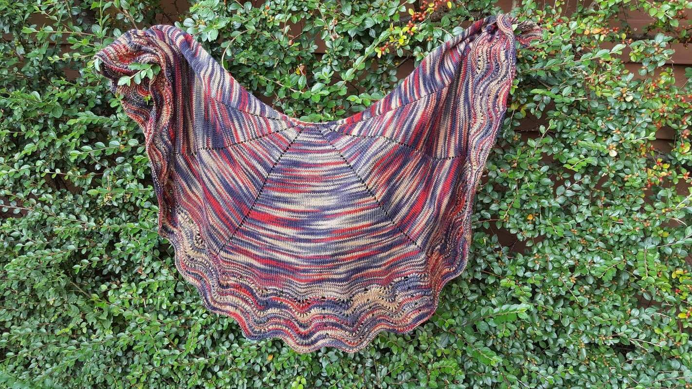 Mehrfarbiges gestricktes Dreieckstuch mit gewelltem Saum vor grünem Blattwerk.