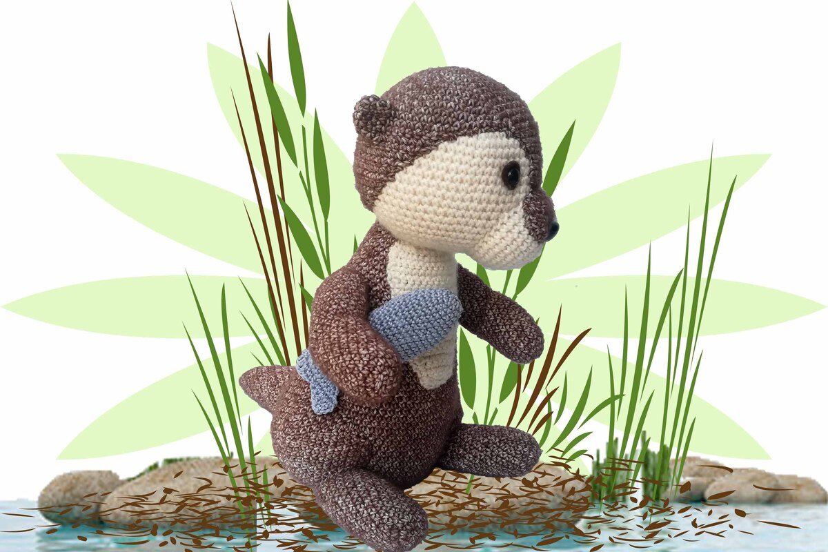 Crocheted brown and cream amigurumi otter holding a small blue fish, seated among reeds