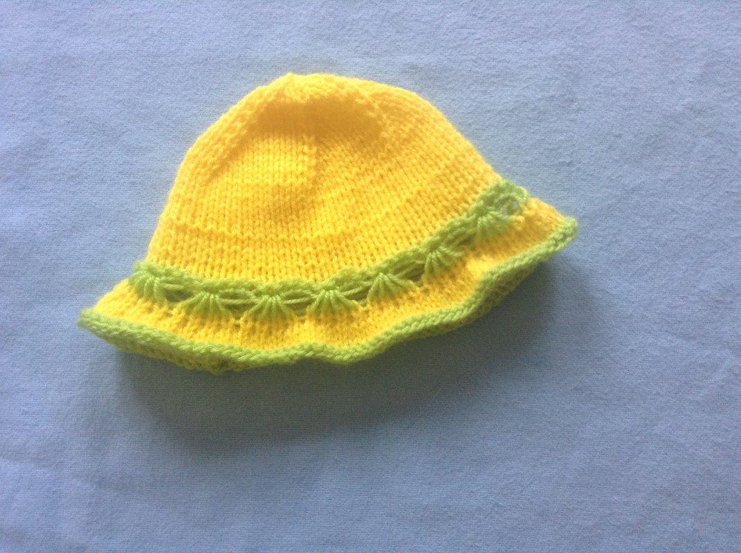 Gestrickter gelber Kinderhut mit grünem Muschelmuster am Rand auf blauem Stoff.