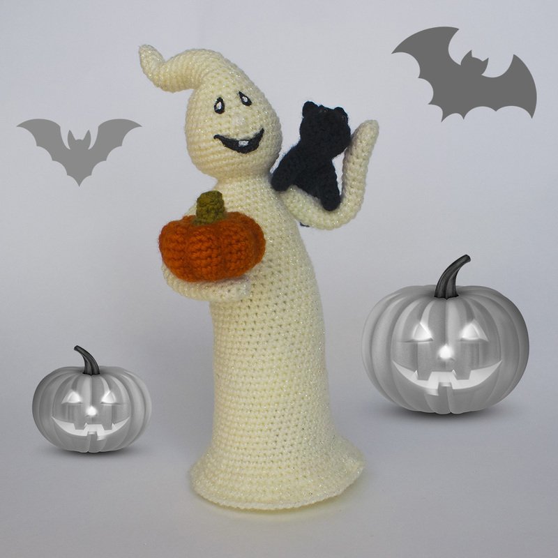 Cream crochet ghost amigurumi holding an orange pumpkin and a small black cat, with bat silhouettes and jack-o'-lantern props