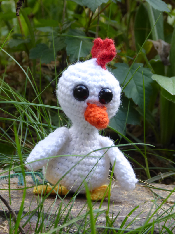 Weißes gehäkeltes Huhn-Amigurumi mit großen schwarzen Augen, rotem Kamm und orangefarbenem Schnabel vor grünem Hintergrund.
