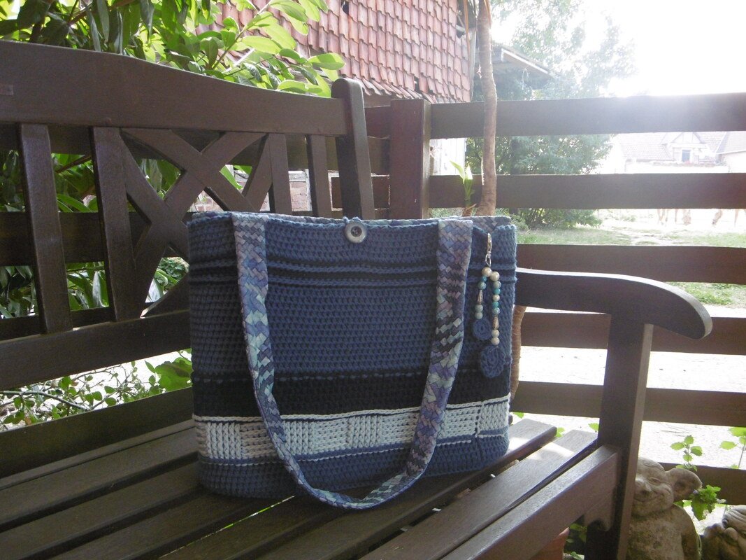 Gehäkelte Umhängetasche in dunklen und hellen Blautönen mit horizontalen Streifen auf einer Holzbank