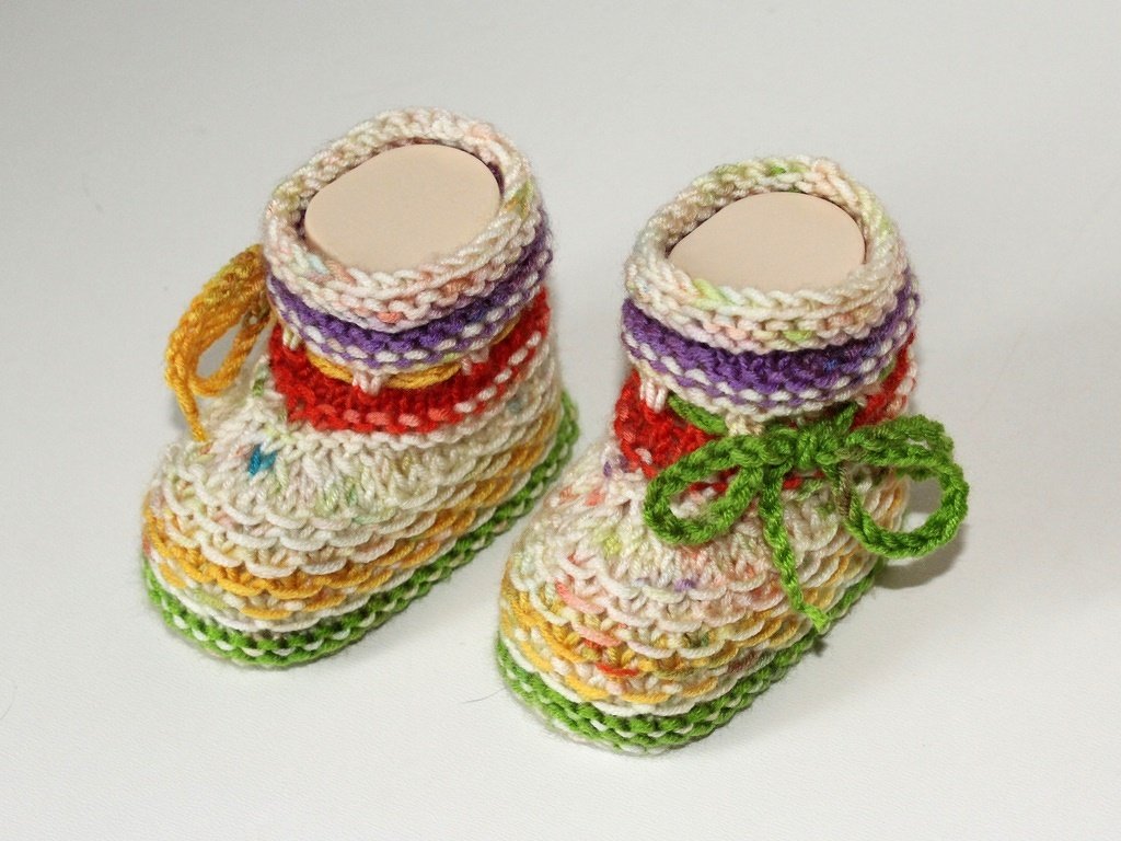 Zwei handgestrickte Babyschuhe mit bunten Streifen, links mit gelber, rechts mit grüner Schleife.