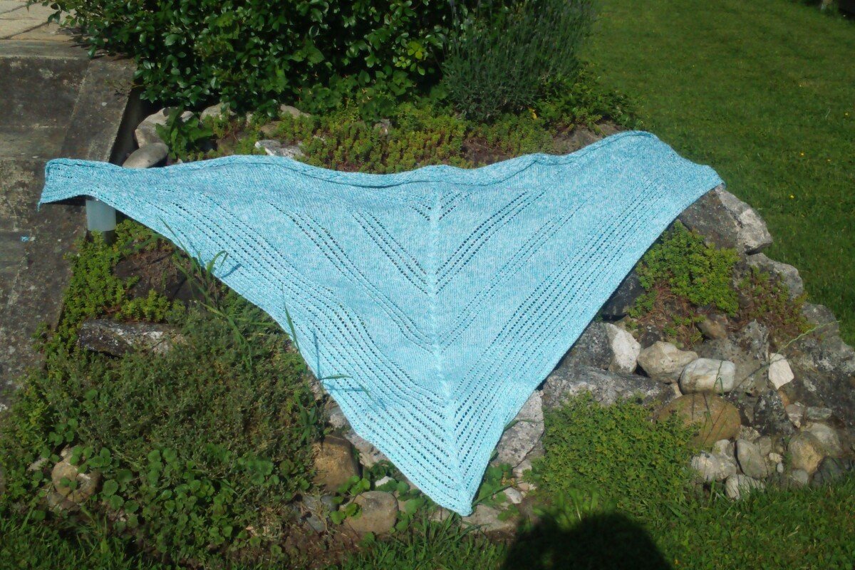 Light blue knitted triangular shawl with eyelet lace rows stretched flat on grass and garden rocks