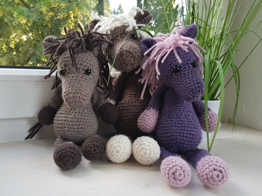 Drei gehäkelte Pony‑Amigurumi mit sichtbaren Mähnen und Sicherheitsaugen auf einer Fensterbank