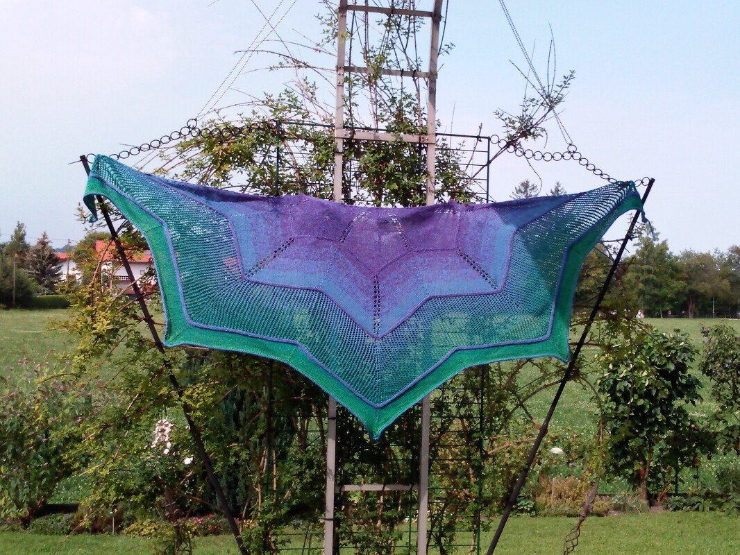Triangular knitted shawl with purple, blue and green gradient displayed outdoors