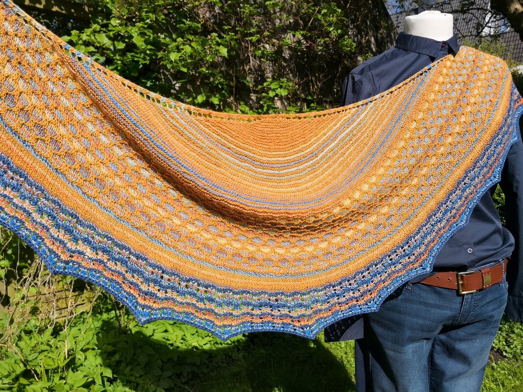Gestricktes halbrundes Tuch in Orange- und Blautönen mit Loch- und Streifenmuster, drapiert an einem Mannequin im Freien.