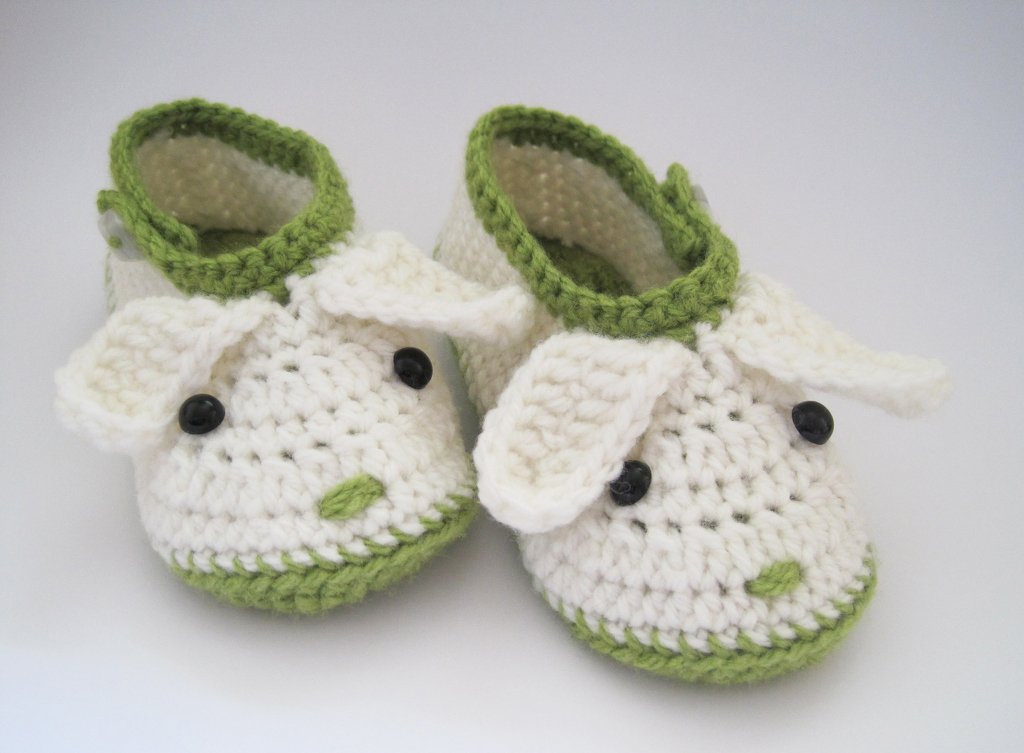 Weiß-grüne handgehäkelte Babyschuhe mit Schäfchendetails, Ohren und dunklen Knopfaugen auf hellem Hintergrund.