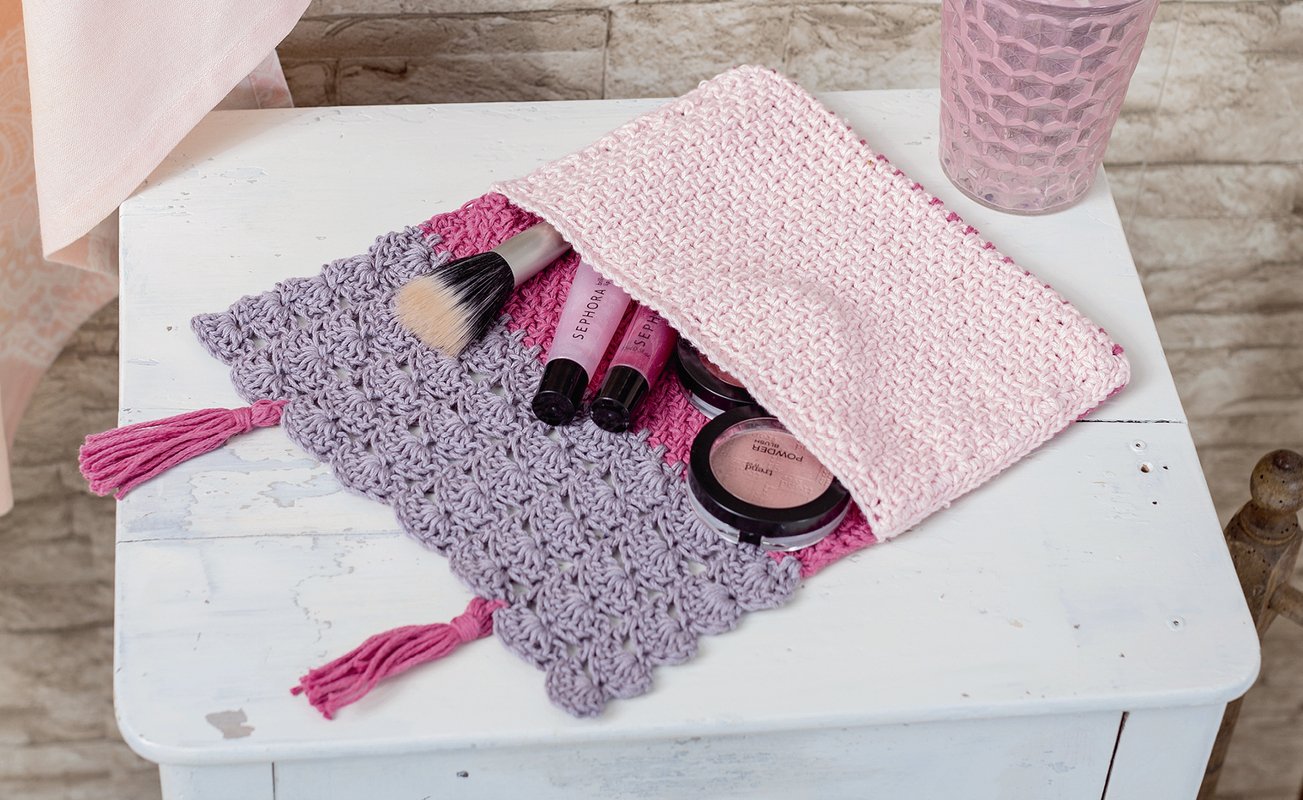 Gehäkeltes pastellfarbenes Täschchen offen mit Make-up-Pinsel, Lipgloss und Kompaktpuder auf weißem Tisch