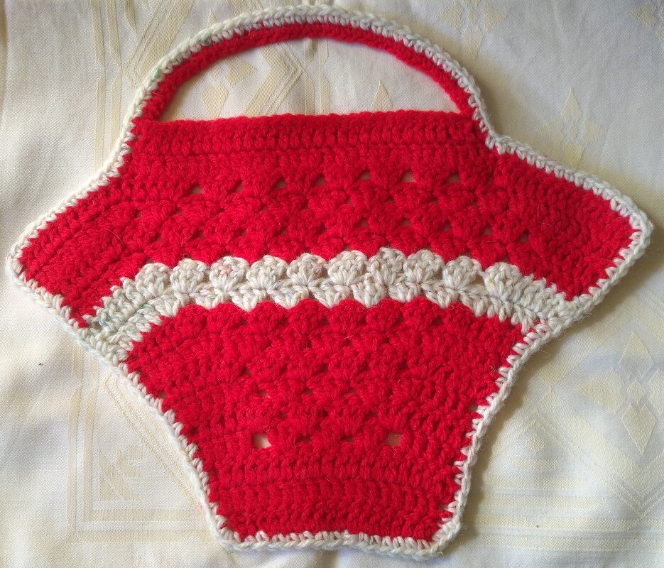 Roter gehäkelter Topflappen in Korbform mit cremefarbener Muschelkante und Henkel auf hellem Stoff.