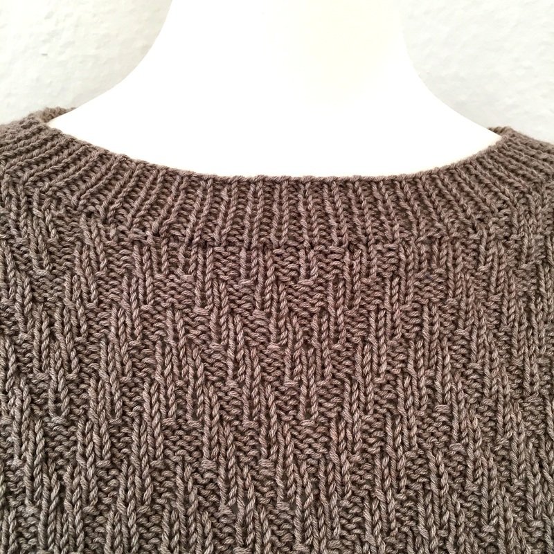 Nahaufnahme des Rundhalsausschnitts und oberen Bereichs eines graubraunen Pullovers mit vertikalem Strukturmuster.
