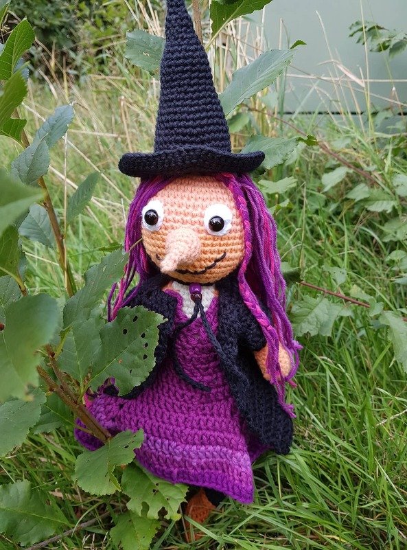 gehäkelte Hexenpuppe mit schwarzem Hut und lila Kleid im Gras