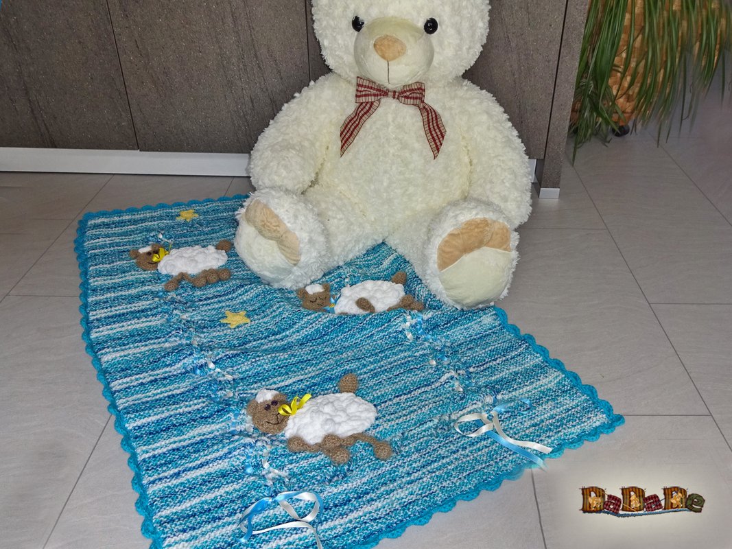 Gestrickte blaue Babydecke mit drei Schäfchen-Applikationen, Zierbändern und gezacktem Rand, darauf ein Teddybär