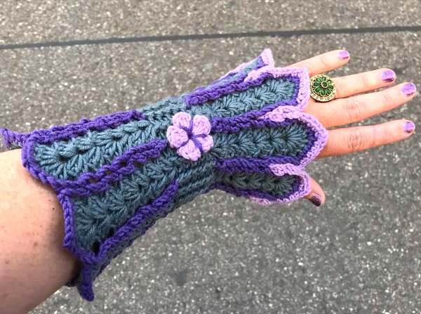 Hand mit gehäkelter petrolfarbener, fingerloser Armstulpe mit violetten Rändern und kleiner Blüte