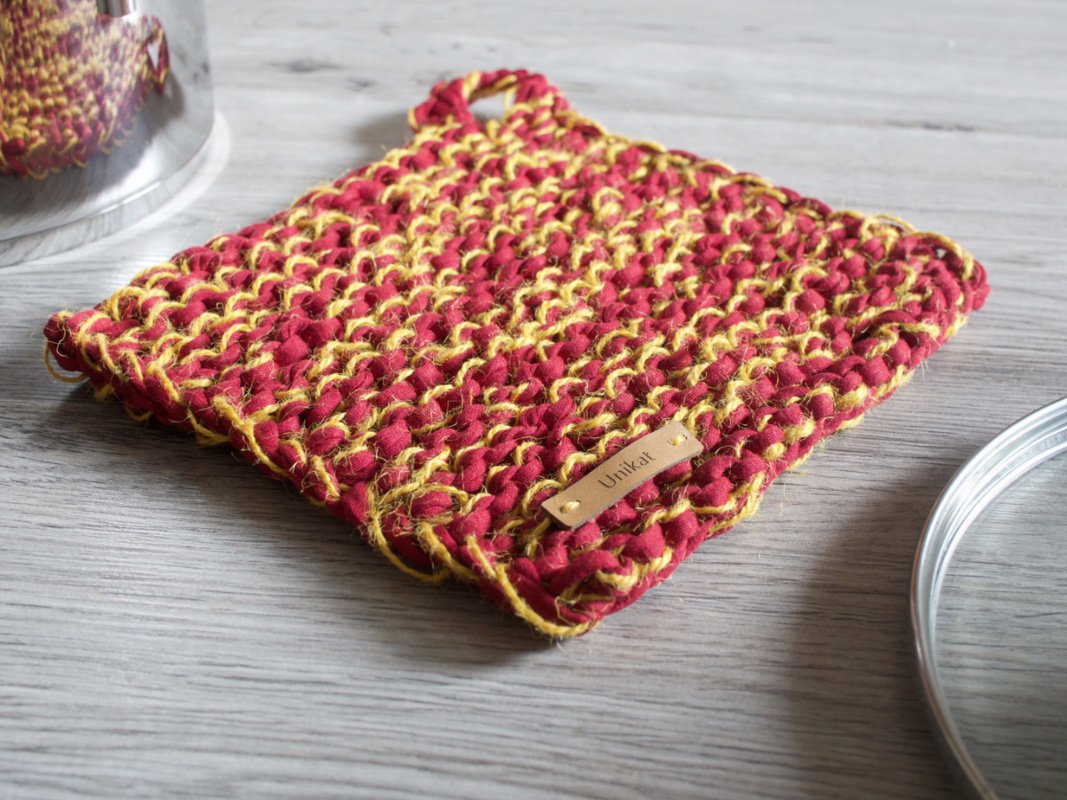 Quadratischer zweifarbiger Topflappen mit Aufhängeschlaufe und Lederetikett auf hellem Holztisch