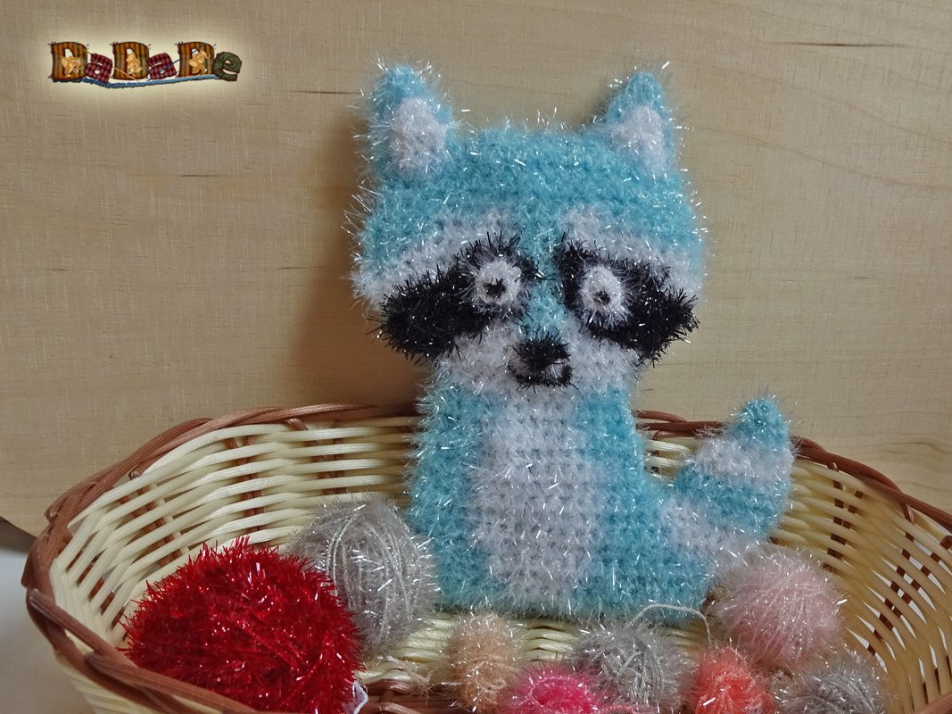 Flauschig gehäkelter blau-weißer Waschbär mit schwarzer Augenmaske im Weidenkorb neben Garnknäueln.