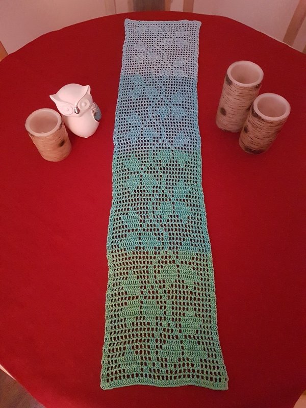 Schmaler gehäkelter Filet-Tischläufer in Blau- und Grüntönen auf rotem Tisch, daneben Keramik-Eule und Kerzenhalter in Holzoptik.