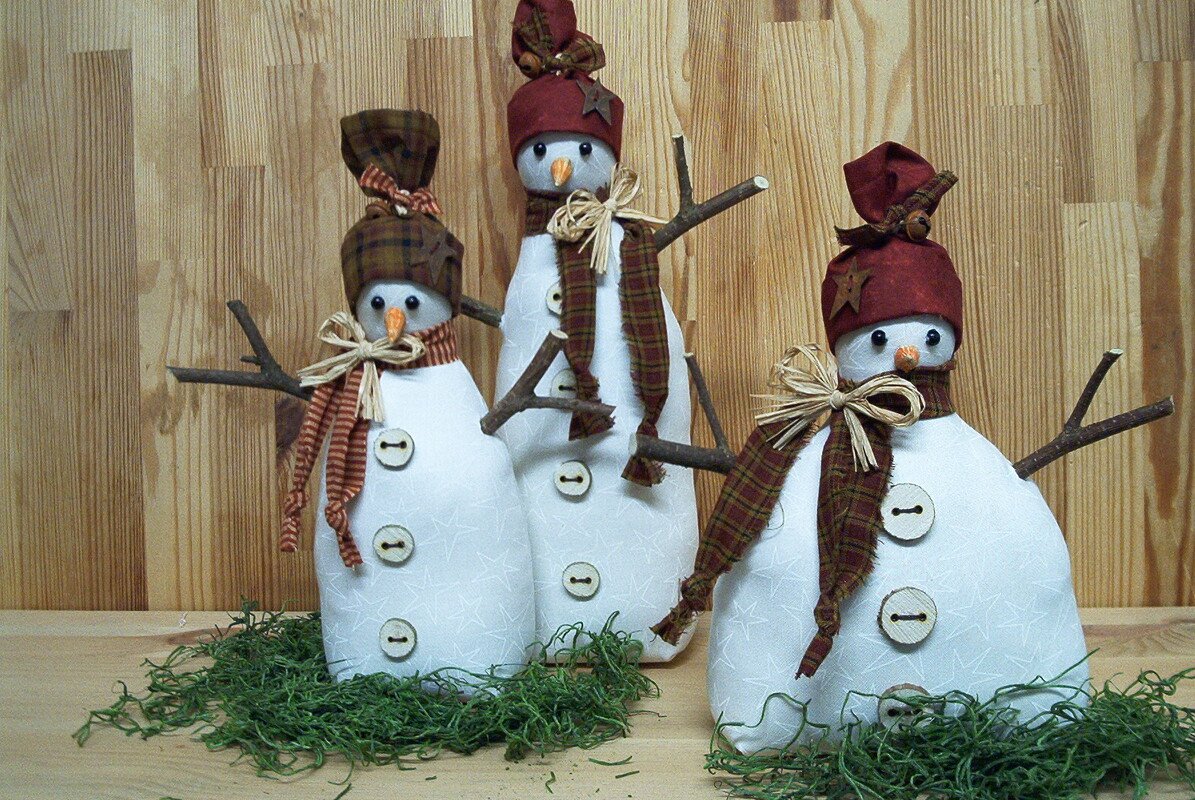 Drei genähte Stoff‑Schneemänner mit Holzknöpfen, karierten Schals, kleinen Mützen und Astarmen