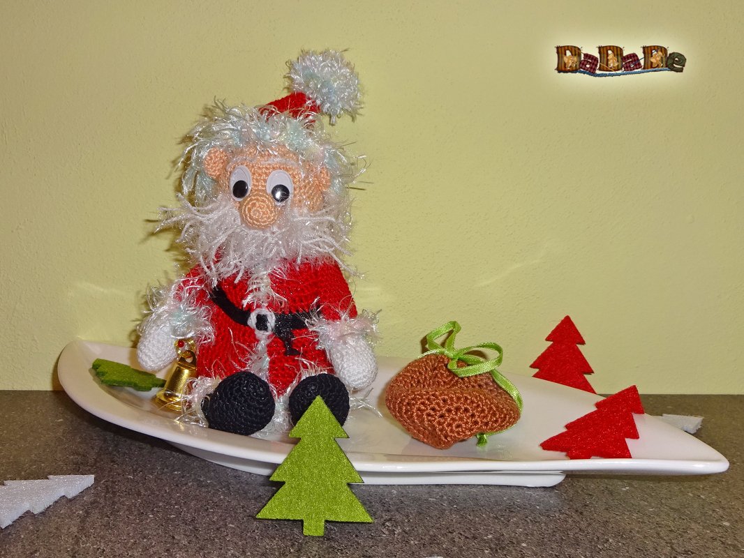 Handgehäkelter Amigurumi-Weihnachtsmann mit weißem Bart und rotem Anzug auf weißer Platte, umgeben von Filz-Tannen, gehäkelter Tasche und goldener Glocke.