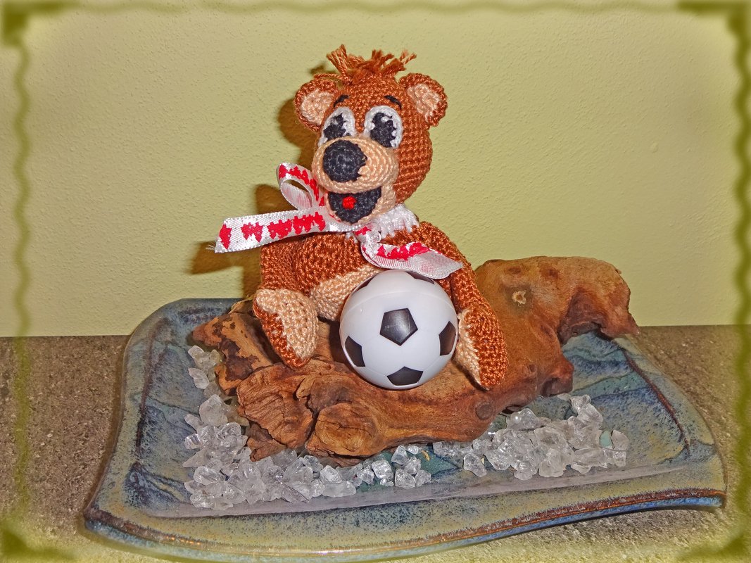 Gehäkelter brauner Teddybär mit rot-weißem Band und kleinem schwarz-weißem Fußball auf Treibholz, Kristallen und blauem Keramikteller