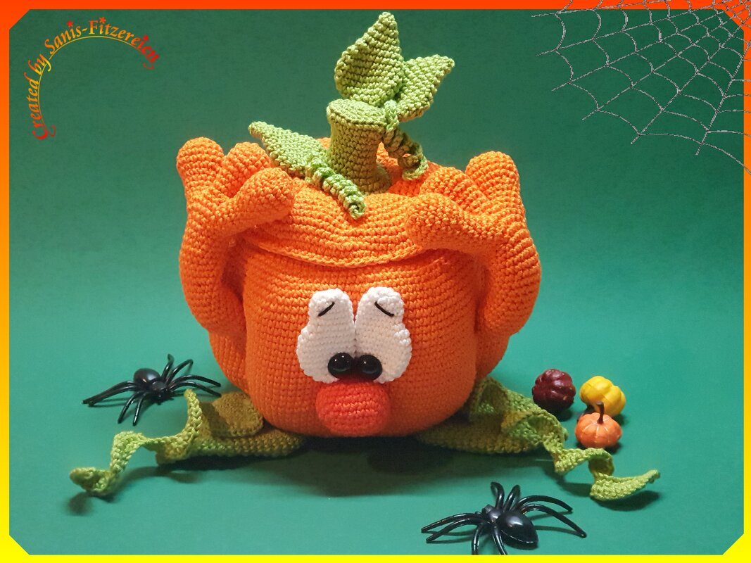 Gehäkelter oranger Kürbis mit weißen Augen, grünen Blättern und kleinen Dekokürbissen vor grünem Hintergrund