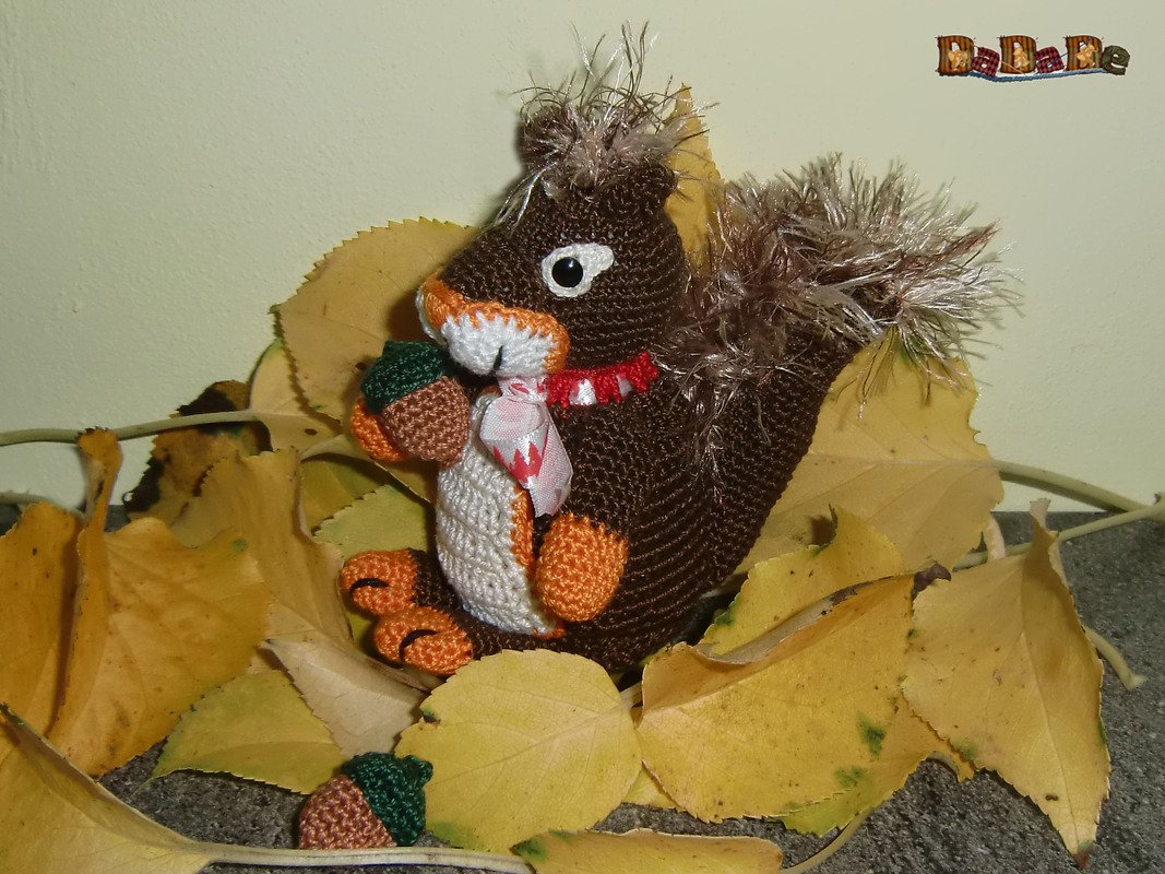 Gehäkeltes braunes Amigurumi-Eichhörnchen mit flauschigem Schwanz hält Eichel auf gelben Blättern