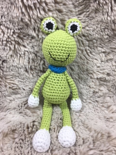 Hellgrüner Amigurumi-Frosch mit blauem Kragen und weißen Händen und Füßen auf flauschigem Hintergrund