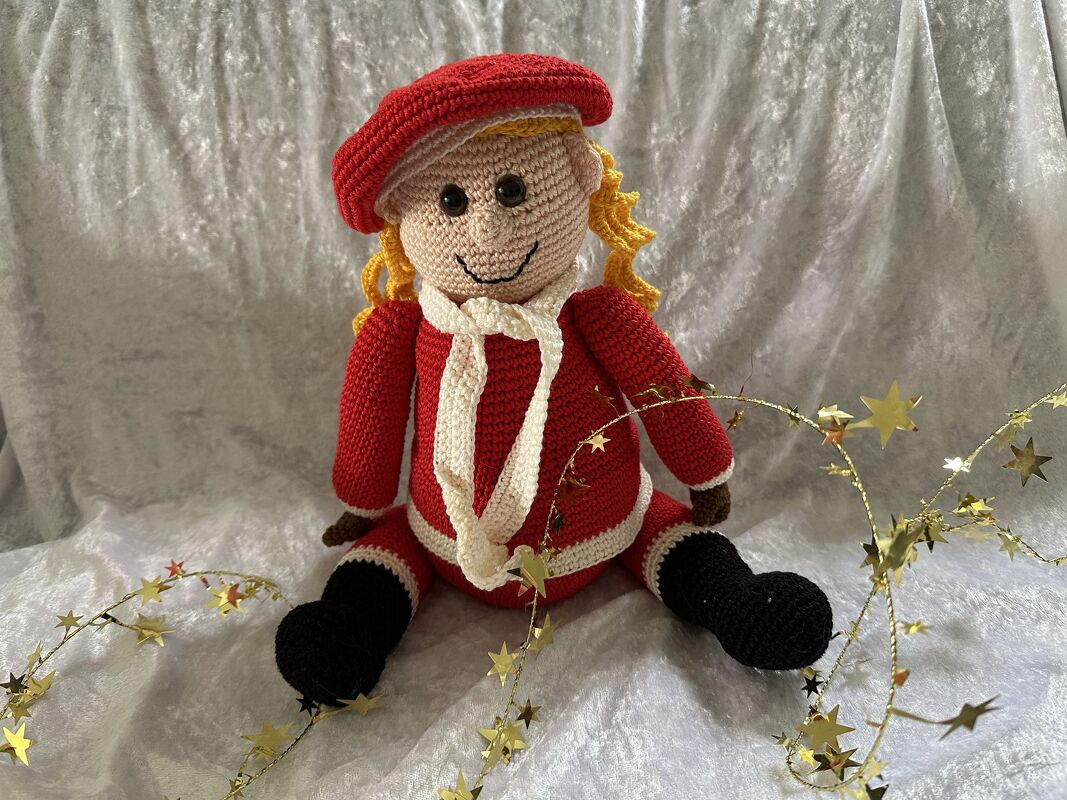 Gehäkelte Amigurumi-Puppe mit roter Mütze, rotem Mantel und langem weißem Schal, gelben Haaren und schwarzen Schuhen, mit Sternengirlande dekoriert