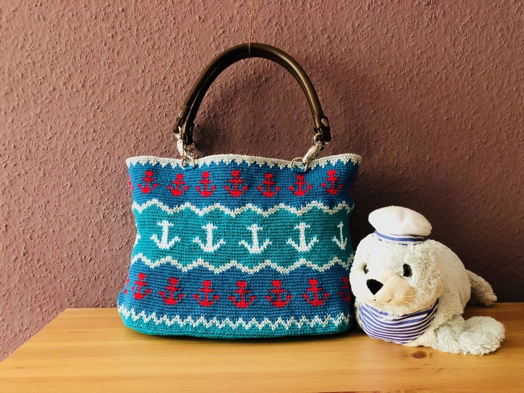 Gehäkelte Henkeltasche blau-türkis mit weißem und roten Anker-Muster, dunkle Henkel, daneben ein Plüschtier