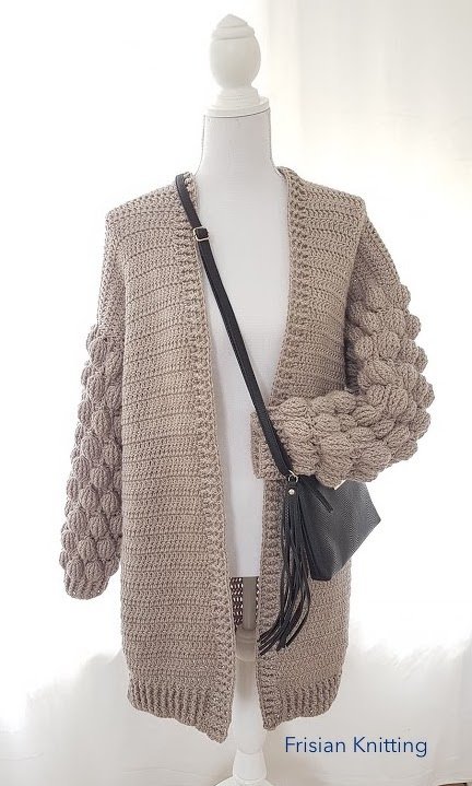 Mannequin wearing a beige open-front crochet cardigan with bubble-stitch sleeves and ribbed hem, styled with a crossbody bag.