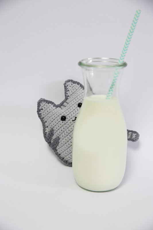 flaches graues Amigurumi-Kätzchen mit dunklem Rand und runden schwarzen Augen, das hinter einer Glasmilchflasche hervorlugt